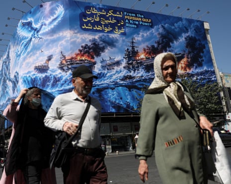 A woman with scarf and a man in a white shirt in foreground, walking past a blue billboard with the words. 'From the PERSIAN GULF, A New Power Rises'.